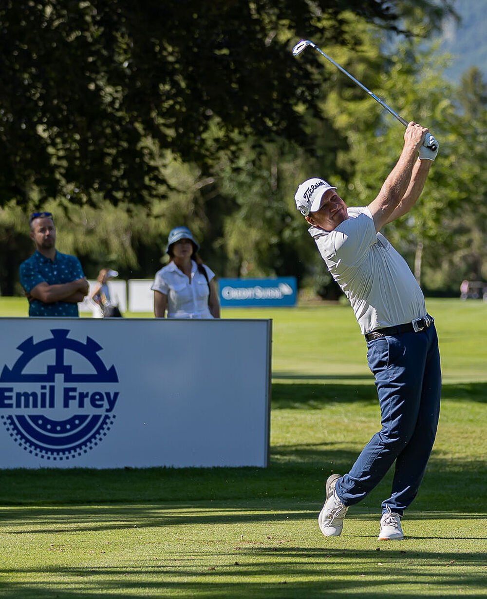 Golfer in der Mitte des Schwungs an einem sonnigen Tag, mit weisser Kappe und blauer Hose. Zwei Zuschauer schauen zu. Bäume und ein Schild im Hintergrund. Ruhige und konzentrierte Stimmung.