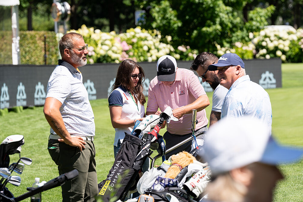 Vier Männer und eine Frau stehen nebeneinander. Einer schaut auf eine Karte, die er unterschreibt. Die Frau hält sie ihm hin. Alle sind mit Golfkleidung bekleidet und stehen auf dem Golfplatz. Im Hintergrund ist eine Webebande zu sehen.