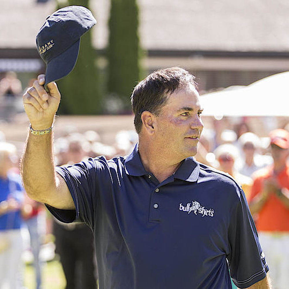 Tim Thelen in einem marineblauen Hemd winkt lächelnd mit einer Baseballmütze, während er auf einem Golfplatz steht. Im Hintergrund klatscht eine farbenfroh gekleidete Menschenmenge enthusiastisch.