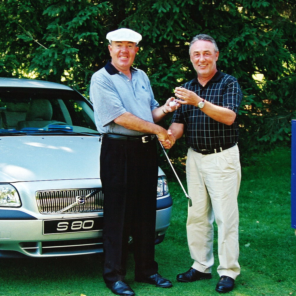 Zwei Männer lächeln und schütteln sich die Hände vor einem silbernen Volvo S80. Ein Mann trägt einen Golfhut und hält einen Schläger. Das Grün im Hintergrund deutet auf eine Parklandschaft hin.