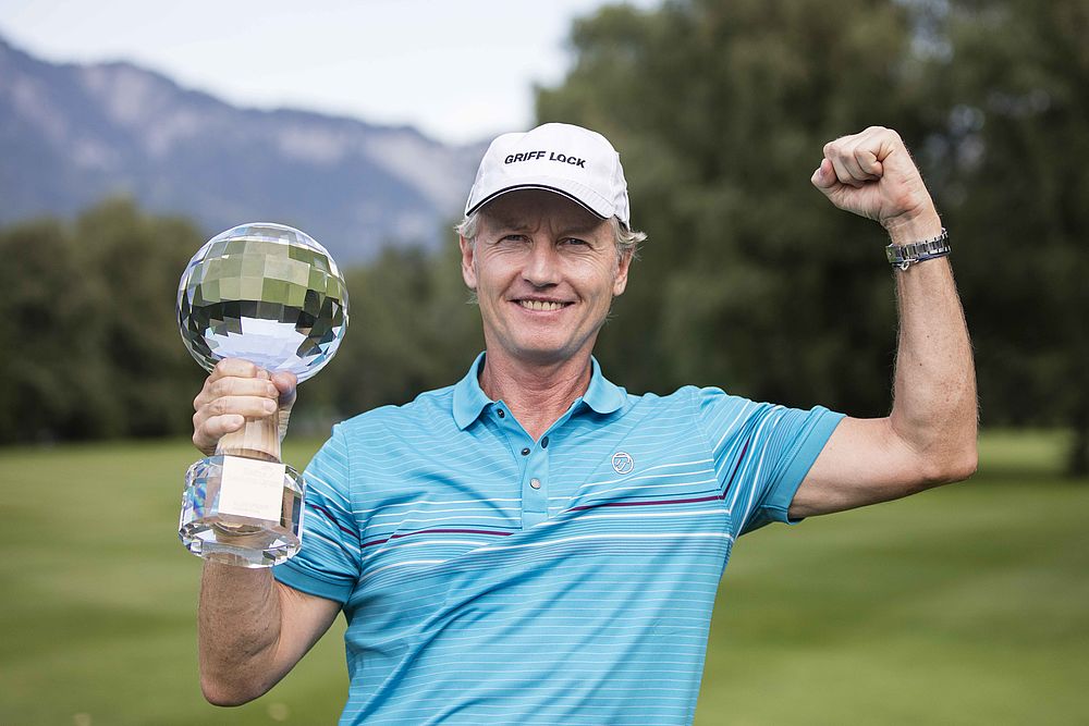 Philip Golding in blauem Polohemd und weißer Mütze hält triumphierend die gläserne Trophäe in der Hand, während er die andere zur Faust hebt. Er steht auf einem Golfplatz mit Bergen im Hintergrund.