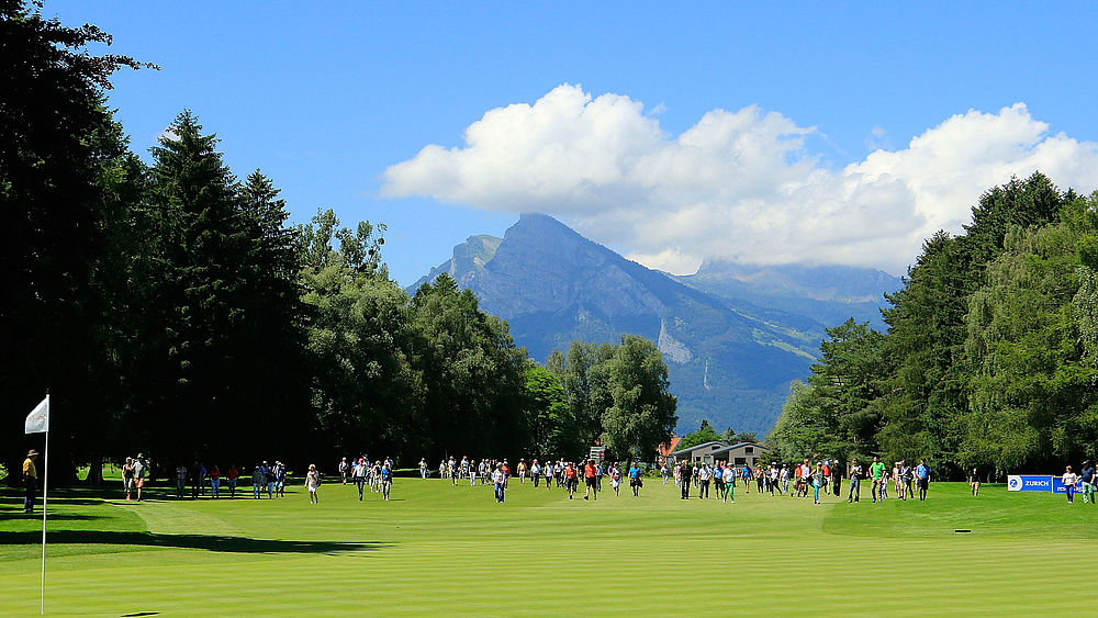 Ein Golfplatz mit einem grünen Feld und verstreuten Bäumen, umgeben von Bergen unter einem klaren blauen Himmel, mit einer entfernten Menschenmenge, die die Szenerie genießt.