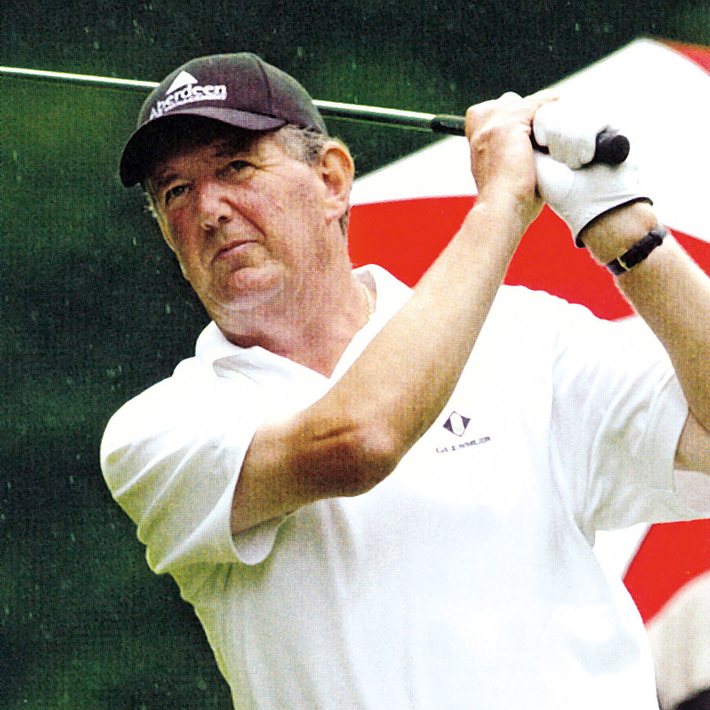 David Huish in weißem Polo und schwarzer Mütze schwingt einen Golfschläger im Freien, konzentrierter Ausdruck. Im Hintergrund sind Bäume und eine rot-weiße Flagge zu sehen, die auf einen Golfplatz hindeuten.