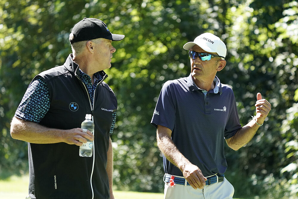 Zwei Männer in Golfkleidung unterhalten sich auf dem Platz, mit Bäumen im Hintergrund. Einer hält eine Wasserflasche. Die Szene vermittelt eine entspannte und konzentrierte Stimmung.