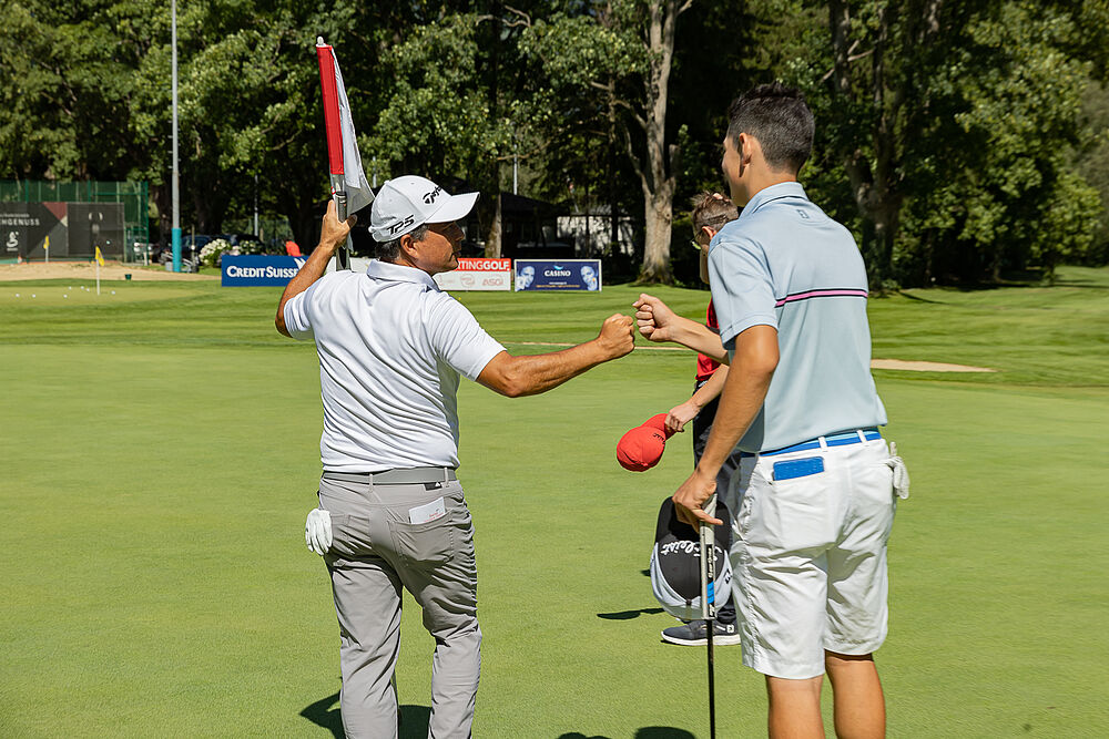 Zwei Golfer tauschen auf einem Putting Green einen Faustschlag aus, um einen erfolgreichen Putt zu feiern. Die Szene spielt an einem sonnigen Tag mit üppigen Bäumen im Hintergrund.
