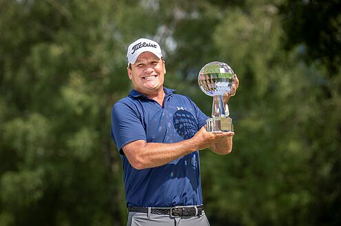 Jarmo Sandelin, der Sieger des Swiss Seniors Open 2024 hält den Glaspokal in die Lüfte. Er trägt ein blaues Hemd und ein weisses Cap und lächelt, während er auf dem Golfplatz die Trophäe hält. Grüne Bäume bilden einen unscharfen Hintergrund.