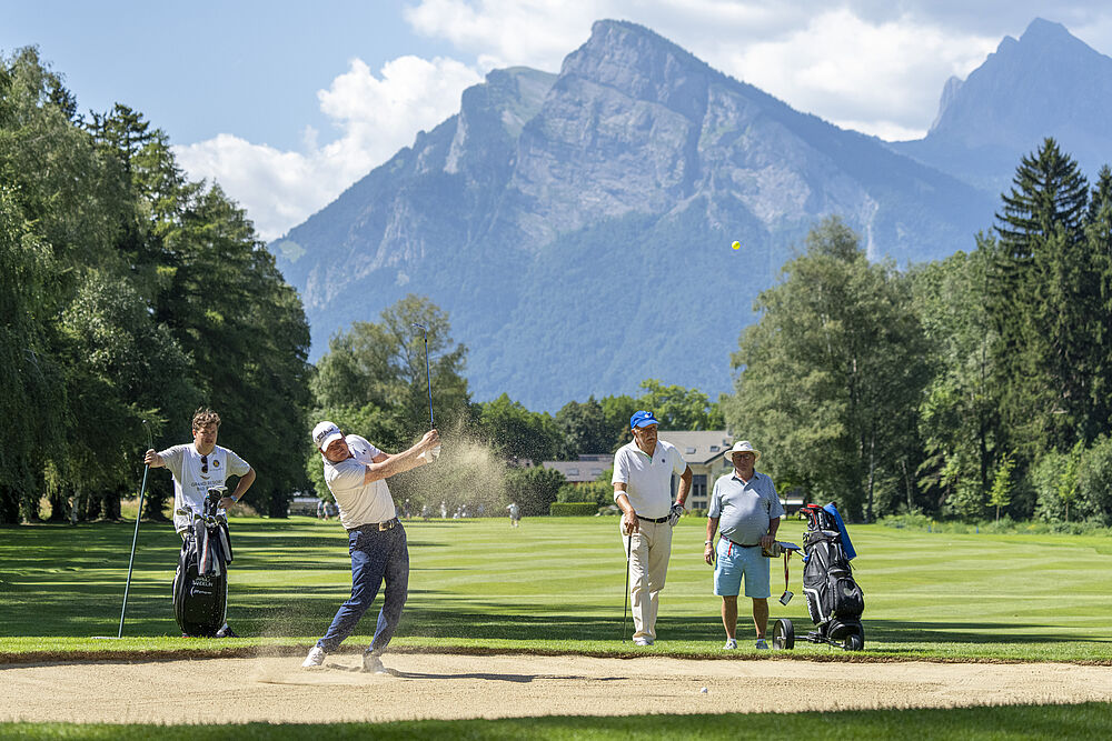 Ein Golfer schwingt in einem Sandbunker auf einem sonnigen Platz, während drei Personen zusehen. Berge und Bäume bilden eine malerische Kulisse und vermitteln Konzentration und Kameradschaft.