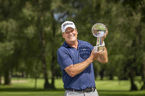 Jarmo Sandelin, der Sieger des Swiss Seniors Open 2024 hält den Glaspokal in die Lüfte. Er trägt ein blaues Hemd und ein weisses Cap und lächelt, während er auf dem Golfplatz die Trophäe hält. Grüne Bäume bilden einen unscharfen Hintergrund.