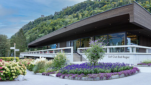 Das Clubhaus des Golf Club Bad Ragaz. Ein modernes Gebäude mit Flachdach, umgeben von Gärten an einem Berghang. Blumen im Vordergrund, Wald im Hintergrund, hell und heiter.
