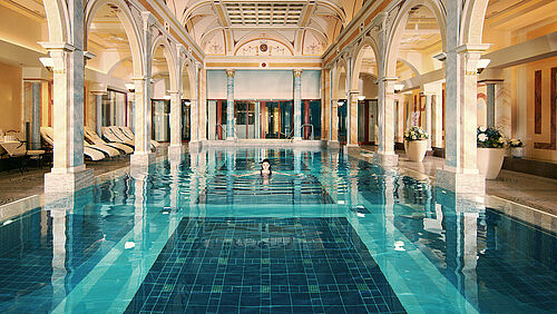 Luxurious indoor pool Helena Bad with elegant arches and a high ceiling. Soft lighting is reflected in the clear blue water and creates a calm, opulent atmosphere.