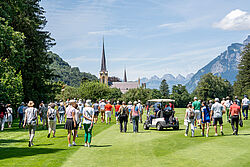 Eine Menschenmenge versammelt sich auf dem Golfplatz bei Loch 12 unter blauem Himmel, umgeben von Bergen und Bäumen. Im Hintergrund ist eine Kirchturmspitze zu sehen, die eine friedliche Atmosphäre schafft.